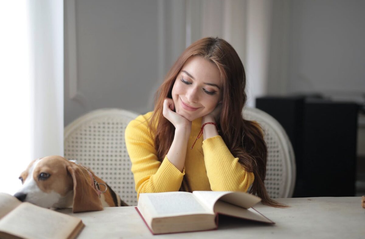 Smiling woman in yellow sweater reading a book with a dog beside her indoors.